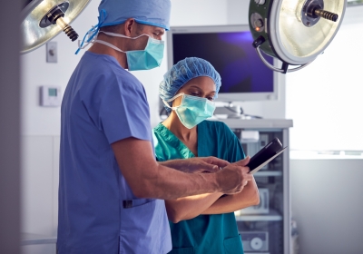 Male And Female Surgeons Wearing Scrubs Looking At Digital Tablet In Hospital Operating Theater